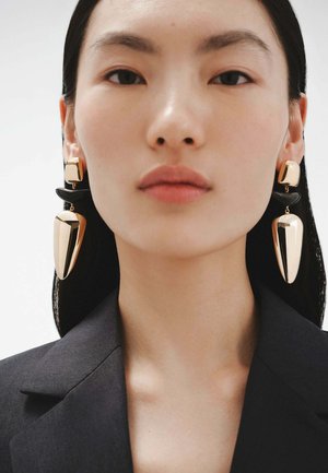 Pendientes abstractos en oro y negro, con formas geométricas y superficies brillantes. La modelo lleva un blazer negro con un escote sencillo.