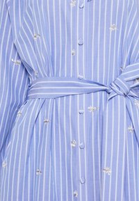 Blårandig skjortklänning med vita vertikala ränder, knäppning fram och midjeband. Har delikat broderi och en lätt tygkvalitet.