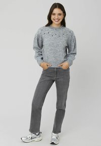 Pull en tricot gris avec un motif texturé et des embellissements au niveau du col, associé à un jean gris et des baskets argentées.