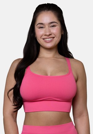 Smiling woman with long dark hair wearing a bright pink ribbed crop top and matching high-waisted bottoms against a plain background.