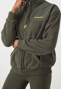 Giacca in pile verde oliva con zip e collo alto, logo Champion in giallo, e cuciture gialle a contrasto su tasche e polsini.