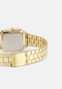 Montre en or avec un boîtier rectangulaire, bracelet en métal à maillons entrelacés et fermoir poli. L'arrière présente une gravure de la marque et une texture lisse.