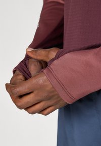 Camiseta de manga larga deportiva en un degradado de burdeos profundo y malva claro, con tejido texturizado y un detalle de orificio para el pulgar en las mangas.