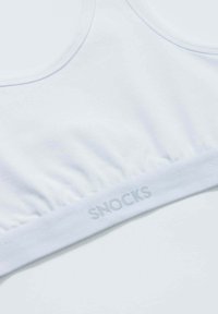 Witte bralette gemaakt van zachte stof met een geplooide band. Heeft een logo-print op de elastische band, wat een subtiele textuur toevoegt.