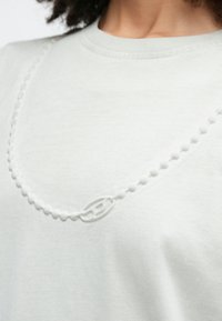 Tuvplāns ar baltu teksturētu T-kreklu, kuram ir pacelts krelles veida ķēdes raksts un neliela cilpa detaļa pie atslēgas kaula.