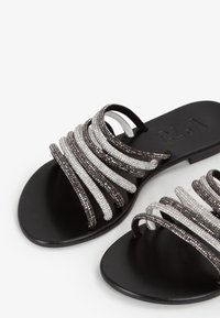 Sandales plates noires avec plusieurs sangles décoratives en tons noirs et argentés alternés, dotées d'une semelle lisse et d'un design minimaliste.