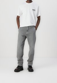 Camiseta blanca de algodón con un logo negro, combinada con jeans grises de corte holgado y botas negras robustas. Estilo casual, ajuste relajado con diseño sencillo.