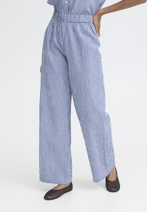 Personne portant un pantalon large à carreaux bleu et blanc et des chaussures plates noires, debout avec une main sur la hanche devant un fond uni.