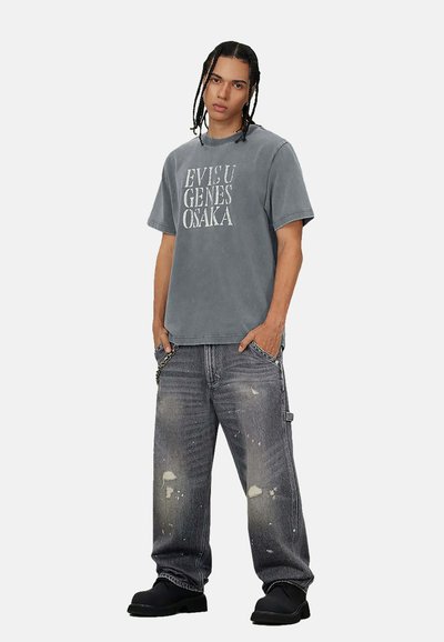 Maglietta grigia oversize con grande testo bianco, abbinata a jeans grigio scuro strappati e scarpe nere. Presenta una vestibilità rilassata e dettagli sottili.