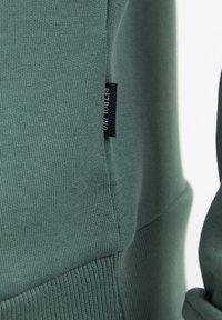 Sudadera verde de mezcla de algodón con textura acanalada, etiqueta en la costura lateral y un ajuste holgado, que enfatiza la simplicidad y la comodidad.