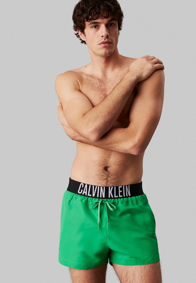 Pantaloni da bagno verdi con una cintura elastica nera che presenta il logo "Calvin Klein". Tessuto liscio, lunghezza sopra il ginocchio, con chiusura a cordoncino.