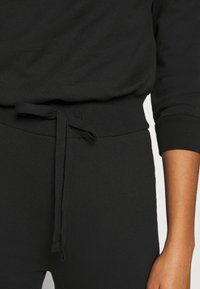 Jersey negro de corte holgado con cintura ajustable mediante cordón. La tela es suave y con textura, presenta un diseño sencillo y contemporáneo.