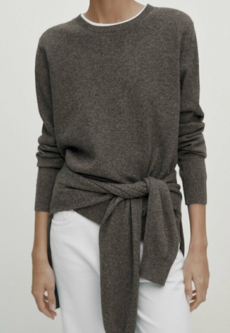 Personne portant un pull marron à manches longues noué à la taille, avec un pantalon blanc, debout devant un fond clair uni.