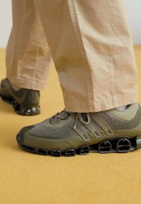 Olive green athletic shoes with mesh panels, ridged soles, and lace-up design. Worn with light beige pants on a yellow background.