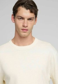 Maglione beige chiaro lavorato a maglia con scollo rotondo, caratterizzato da un logo sottile sul petto e una finitura morbida e testurizzata.