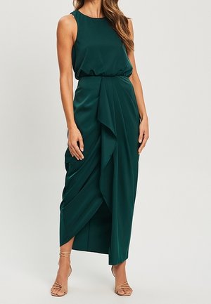 Ärmelloses, dunkelgrünes Midikleid mit tailliertem Schnitt und mehrlagigem, drapiertem Rock, getragen mit beigen Riemchenabsätzen.