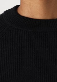 Schwarzer gerippter Pullover mit Rundhalsausschnitt und strukturiertem Stoff. Nahaufnahme zeigt das Strickmuster und die glatte Oberfläche.