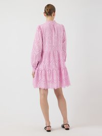 Ružičasta pletačka haljina s dugim rukavima, valovitim rubom i uzorkom otvorenog rada. Ima visoki izrez i suknju u slojevima.