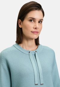 Pull bleu clair avec texture côtelée horizontale, col rond et cordons de serrage ajustables avec embouts en métal. Tissu doux, coupe décontractée.