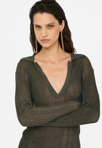 Stricktop mit tiefem V-Ausschnitt in Dunkelgrün und geripptem Muster. Verfügt über lange Ärmel und einen subtilen Schimmer. Das Model posiert mit verschränkten Armen.
