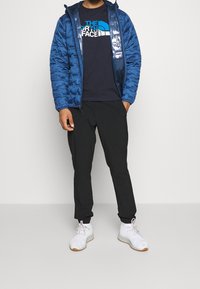 Chaqueta de plumas aislante azul con un acabado brillante, llevada sobre una camiseta negra, combinada con pantalones negros y zapatillas deportivas blancas.