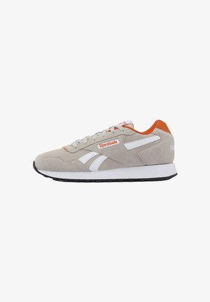 Lichtgrijze suède Reebok-sneaker met witte strepen, oranje voering en zwarte zool, zijaanzicht op een witte achtergrond.