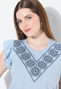 Camiseta azul claro con mangas cortas acampanadas, que presenta patrones circulares bordados en negro en un escote en forma de V. Textura de tejido suave.