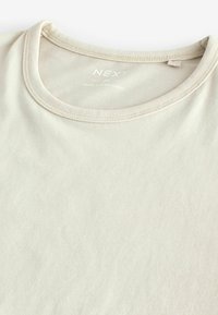 Tricou bej deschis cu guler rotund, material moale și cu o cusătură la guler. Eticheta spune "NEXT" împreună cu informații de îngrijire.