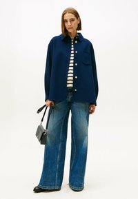 Marineblauwe oversized wollen blazer met gouden knopen, gedragen over een gestreept shirt, gecombineerd met denim jeans met wijde pijpen en een zwarte handtas.