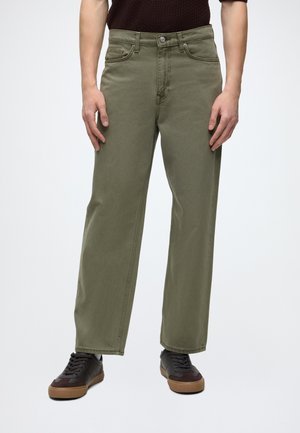 Hombre con pantalones rectos color verde oliva, camisa marrón oscuro y zapatillas oscuras con suelas de goma, de pie frente a un fondo claro y liso.