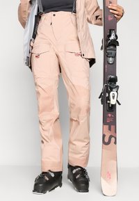 Calças de esqui rosas com múltiplos bolsos, alças ajustáveis, textura suave; usadas com botas de esqui pretas e segurando um esqui com ligações.