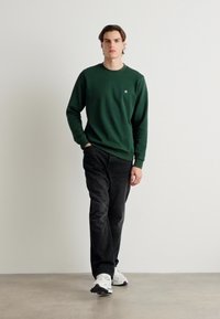 Pullover verde a collo rotondo, pantaloni neri e sneaker bianche. Il pullover presenta un logo discreto sul petto; i pantaloni sono in velluto a coste.