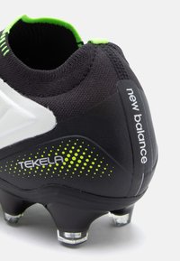 Mustandid jalatsid, mis on musta värvi ja millel on tekstureeritud pealispind, valged aktsendid ja rohelise täppidega muster. Kaubamärgi "New Balance" ja "Tekela" logo on nähtavad küljel.