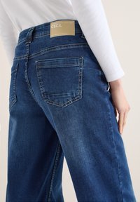 Personne portant un jean en denim bleu foncé avec des poches arrière et une étiquette en cuir beige portant l'inscription "CECIL" sur la ceinture, associée à un haut blanc à manches longues.