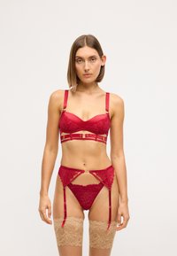 Rotes Spitzen-BH- und Strumpfhalter-Set mit Satin-Trägern, goldenen Hardware-Details, filigraner Spitzenverzierung und durchsichtigen beige Oberschenkelstrümpfen.