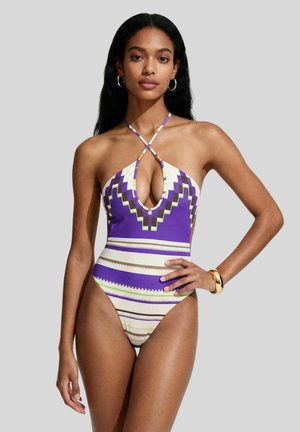 Donna con lunghi capelli neri che indossa un costume da bagno a collo all'americana con motivo geometrico viola, bianco e verde, in piedi con una mano sul fianco.