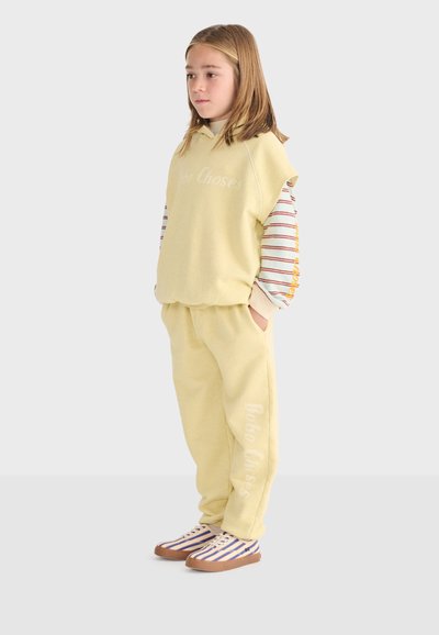 Enfant debout portant un sweat à capuche sans manches jaune et un pantalon, un t-shirt à manches longues rayé et des baskets rayées sur fond uni.
