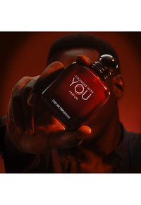 Mano che tiene una bottiglia rossa di profumo Emporio Armani "Stronger With You" su sfondo arancione scuro.