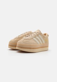 Beige suéd platform tornacipő három fehér csíkkal, kerek orral, lapos fűzőkkel és világosbarna gum talppal és középtalppal. Arany színű logó a oldalon.