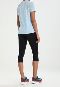 Personne debout portant une chemise à manches courtes bleu clair, des leggings capri noirs et des chaussures de course grises avec des accents rouges, de dos sur fond blanc.