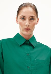Grüne Hemdbluse mit strukturiertem Kragen, einer Brusttasche und einer glatten, leicht glänzenden Textur.