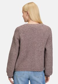 Kobieta z blond włosami, ubrana w strukturę sweter w kolorze mauve i jasnoniebieskie dżinsy, stoi tyłem na białym tle.