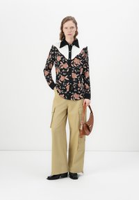 Sorte skjorte med blomstermønster og hvit krage, kombinert med beige vide cargo-bukser og en brun veske. Modellen står mot en hvit bakgrunn.