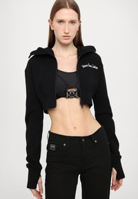 Melnā īsā zip-up kapuce ar kapuci, ar izšūtu logo. Uzvilkta virs melnā krūštura un kombinēta ar melnām piegulošām biksēm.