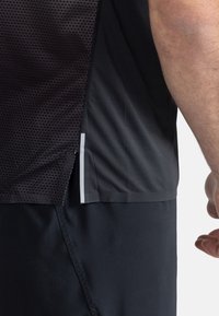Detailní záběr na osobu v černém síťovaném sportovním topu a černých kalhotách, ukazující boční šev s malým reflexním pruhem.