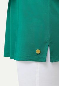 Camicetta verde a maniche corte con tessuto testurizzato, fessure laterali e un'etichetta circolare dorata vicino all'orlo, abbinata a pantaloni bianchi.