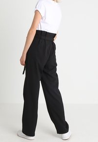 Pantalons noir à jambe large avec une taille haute, ceinture en tissu et finition texturée, associés à une chemise blanche à manches courtes et des baskets blanches.
