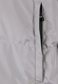 Grå jakke stof med en lynlåslomme detalje. Glat tekstur og synlige syninger understreger funktionalitet og design.