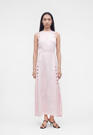 Femme debout dans une robe sans manches rose pâle longueur cheville avec une bordure en dentelle et portant des sandales à talons ouvertes noires.