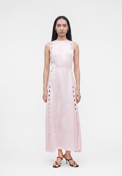 Femme debout dans une robe sans manches rose pâle longueur cheville avec une bordure en dentelle et portant des sandales à talons ouvertes noires.
